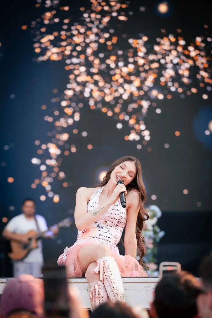 Una cantante vestida de rosa actúa en el escenario con un micrófono, sentada en el borde del escenario. Al fondo se ven unas luces desenfocadas y, a lo lejos, un guitarrista tocando. En primer plano se ve al público.