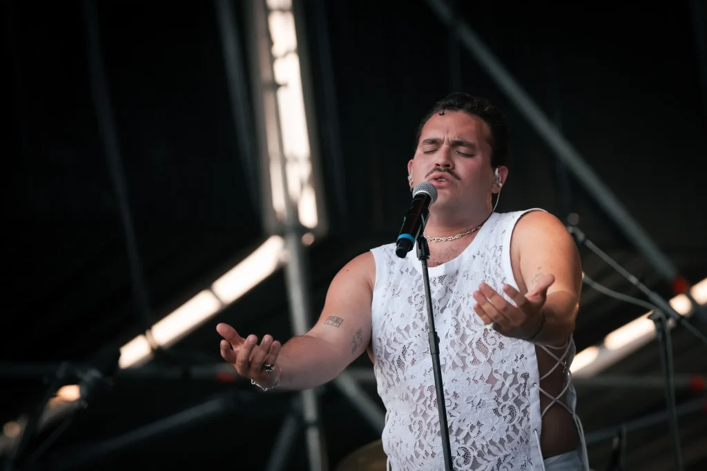 Una persona que lleva un top blanco de encaje sin mangas canta ante un micrófono en un escenario al aire libre, con los ojos cerrados y las manos levantadas. Al fondo se ven el equipo y las estructuras del escenario.