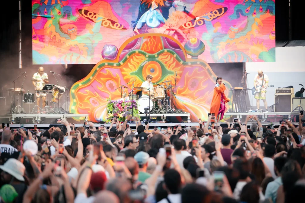 Una banda actúa en un escenario al aire libre decorado con motivos psicodélicos de colores vivos, mientras una gran multitud observa y hace fotos. El escenario cuenta con fondos de colores y músicos que tocan en directo.