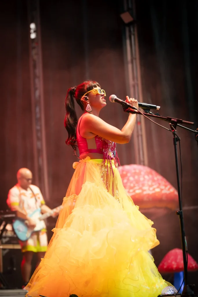 Una cantante, vestida con un vestido de colores amarillo brillante y rosa y con gafas de sol amarillas, actúa en el escenario, sosteniendo un micrófono. Al fondo se ve a un guitarrista, y en el escenario hay grandes setas de atrezo.