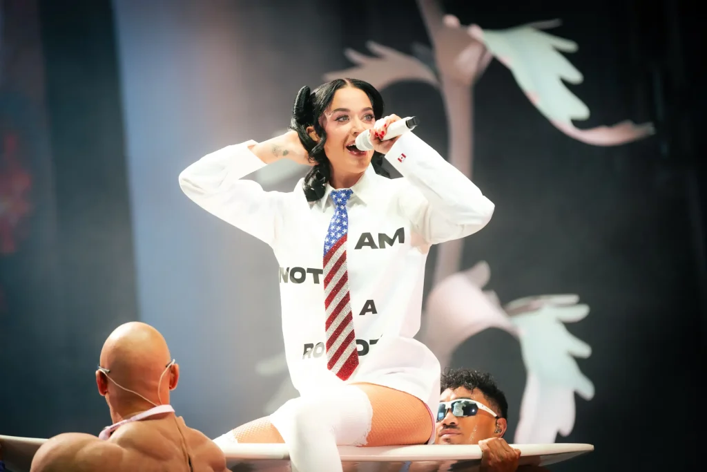 Una mujer con una camiseta blanca en la que se lee «I AM NOT A ROBOT» y una corbata con la bandera estadounidense canta por un micrófono mientras está sentada sobre una tabla de surf sostenida por dos hombres en el escenario. El fondo está decorado con formas blancas estilizadas.