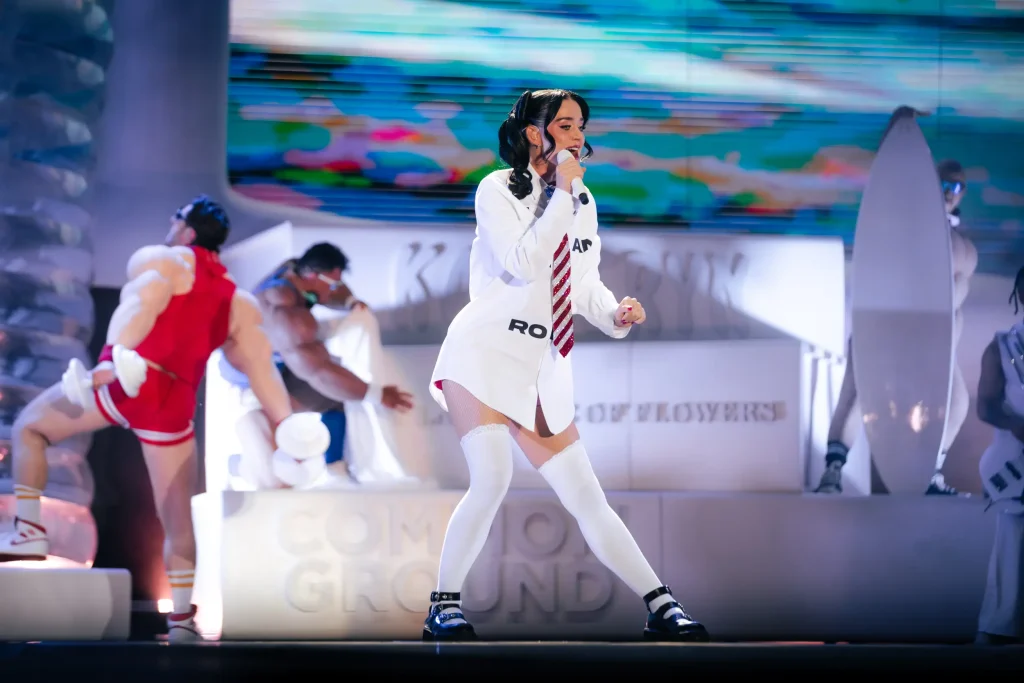 Una mujer con camisa blanca, corbata y calcetines hasta la rodilla canta ante un micrófono en el escenario, rodeada de bailarines con ropa deportiva y con un fondo colorido y abstracto a sus espaldas.