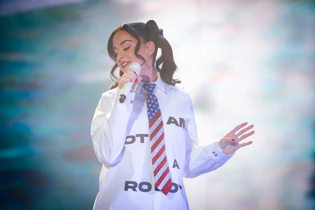 Una mujer de pelo oscuro recogido en coletas canta ante un micrófono. Lleva una camisa blanca abotonada con una corbata brillante con la bandera estadounidense y las letras «AM» y «ROLD» impresas en ella.