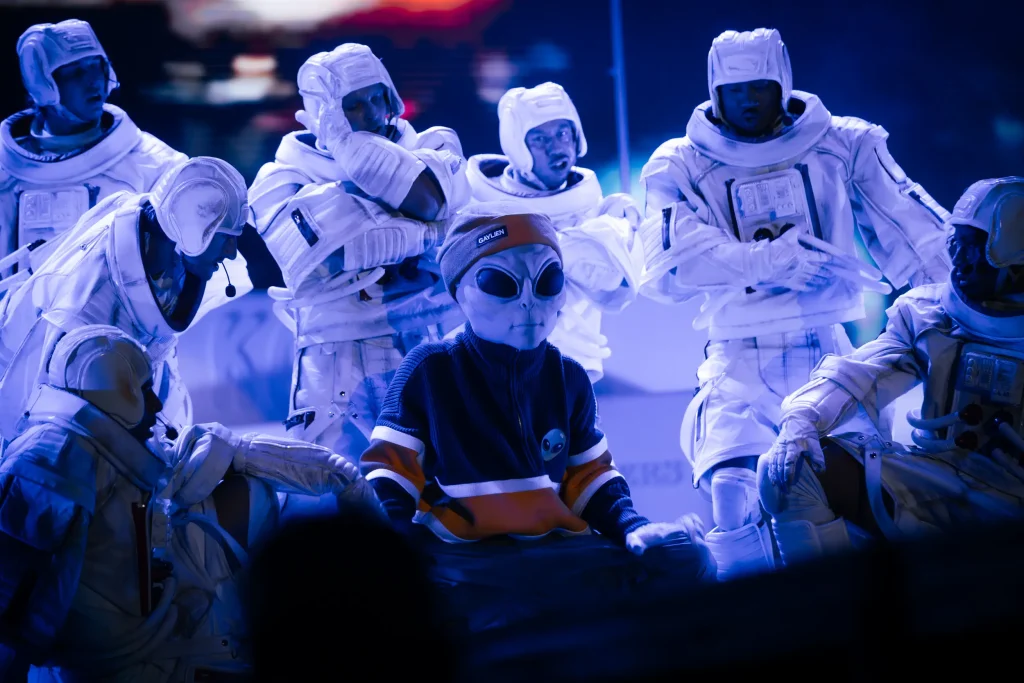 Un grupo de personas con trajes blancos de astronauta rodea a una persona que lleva una máscara gris de extraterrestre, gafas de sol y un jersey a rayas rojas y naranjas, todo ello bajo la iluminación azul del escenario.
