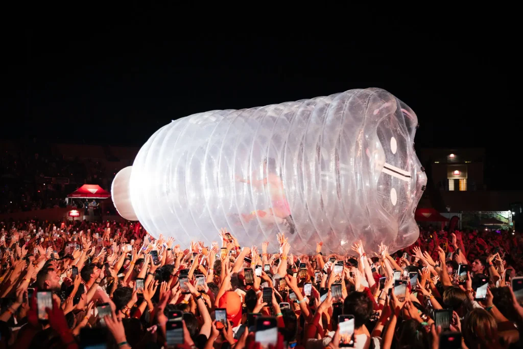 Una gran estructura hinchable transparente con forma de botella de plástico está siendo transportada por encima de la multitud en un evento nocturno, mientras muchas personas alzan los brazos y se hacen fotos con sus móviles.