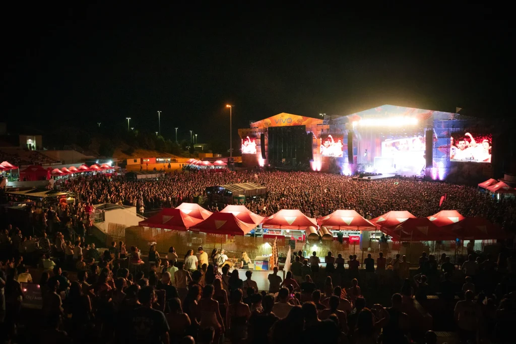 Una gran multitud se congrega en un concierto al aire libre por la noche, con dos escenarios intensamente iluminados. En primer plano se alinean hileras de puestos de venta de colores rojo y blanco, y varias pantallas gigantes muestran primeros planos de los artistas en el escenario.
