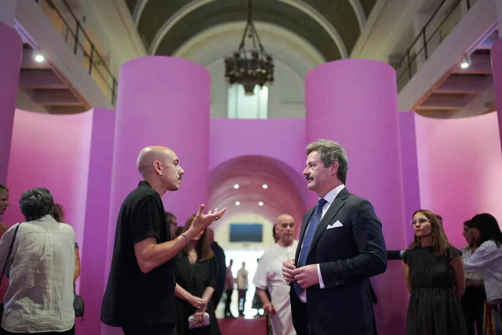 Dos hombres están de pie charlando frente a unas grandes columnas rosas en el interior de la Sala Alcalá 31, un escenario que recuerda a La Movida madrileña. Varias personas más se mezclan entre el público al fondo, lo que sugiere que se trata de un evento o una exposición en el interior en la que se muestran obras únicas.