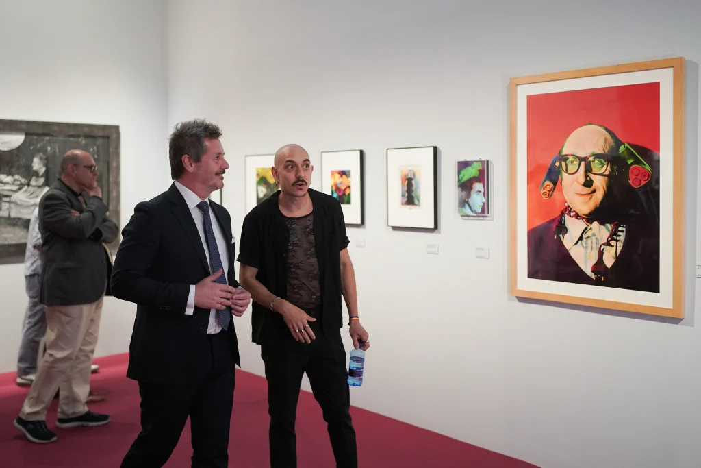 Dos hombres se encuentran en la Sala Alcalá 31, admirando un retrato lleno de color; otros visitantes contemplan las obras que se ven al fondo. La moqueta roja y las paredes blancas de la galería están adornadas con obras enmarcadas, que reflejan el espíritu vibrante de La Movida madrileña.