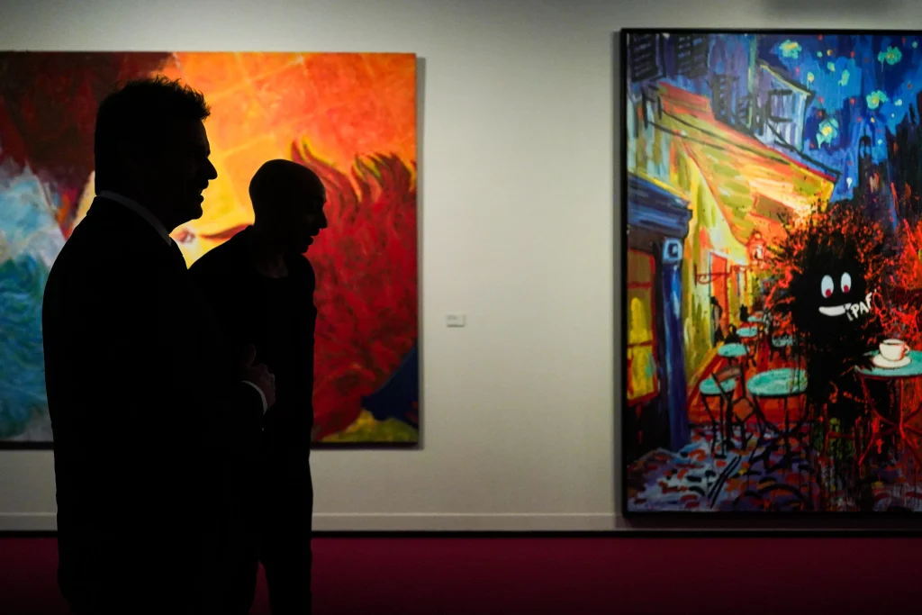 Dos personas, vistas de perfil, se encuentran frente a coloridas pinturas abstractas —obras inspiradas en La Movida madrileña— en una galería de arte con moqueta roja y paredes blancas, que recuerda a la animada Sala Alcalá 31.