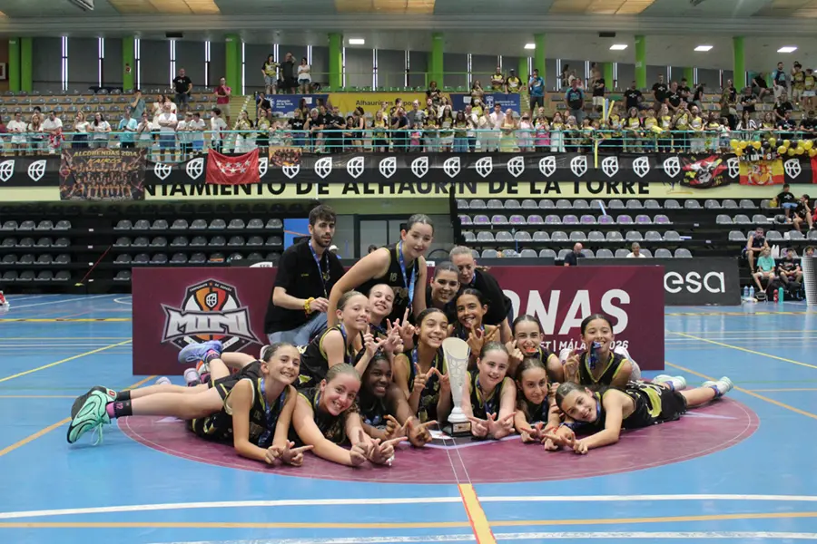 El equipo femenino de baloncesto de Uros de Rivas posa tumbado en el suelo del pabellón, delante de un cartel, sonriendo a la cámara. Los espectadores están sentados en las gradas detrás de ellas, y de la barandilla cuelgan pancartas que celebran los logros de su cantera. La escena se desarrolla en un recinto cubierto con una iluminación intensa, lo que refleja la emoción por su reciente victoria en el título nacional.