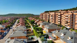 Vista aérea de un barrio residencial con hileras de casas, céspedes verdes y un edificio de apartamentos alto a la derecha. Al fondo se divisan colinas y un cielo azul claro. La zona destaca por su cuidada planificación urbanística, con bonificaciones especiales en el IBI disponibles para las familias numerosas de la comunidad.
