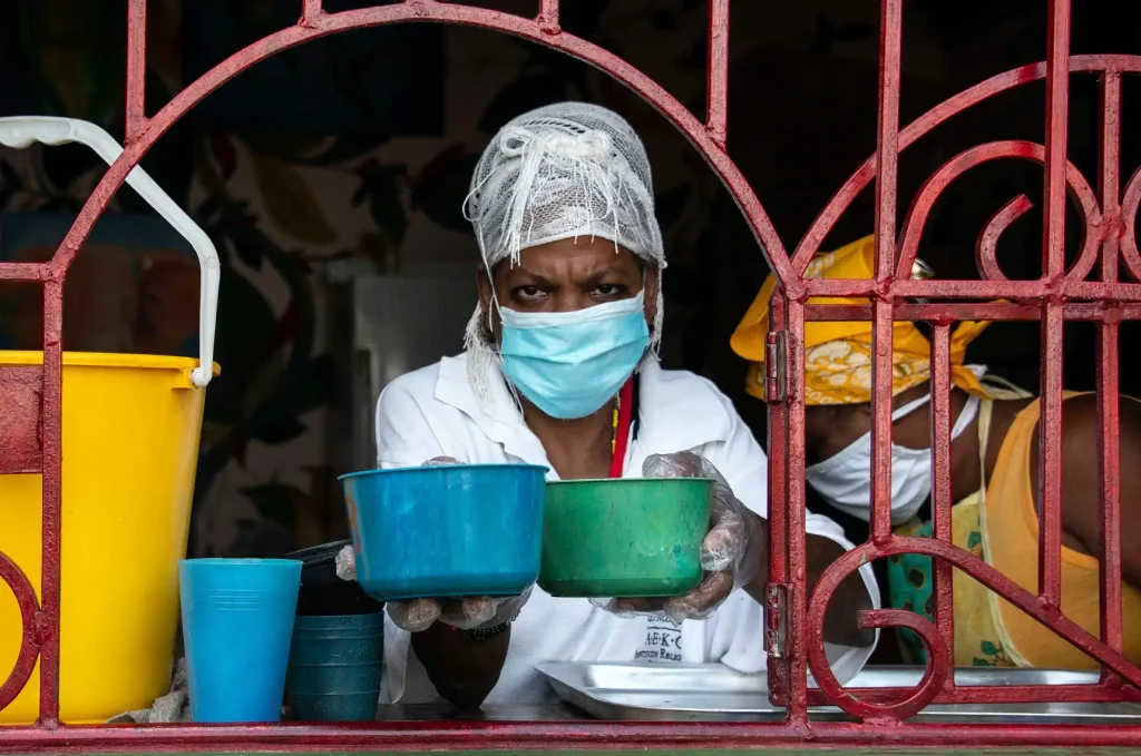 Una persona que lleva mascarilla, redecilla y guantes entrega dos cuencos de plástico a través de una ventana enrejada, como parte de una campaña para distribuir legumbres en Cuba. Junto a ella se encuentra otra persona con un pañuelo amarillo en la cabeza. A la izquierda se ve un cubo amarillo.