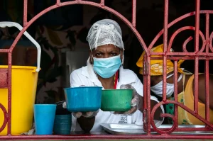 Una persona que lleva mascarilla, redecilla y guantes entrega dos cuencos de plástico a través de una ventana enrejada, como parte de una campaña para distribuir legumbres en Cuba. Junto a ella se encuentra otra persona con un pañuelo amarillo en la cabeza. A la izquierda se ve un cubo amarillo.