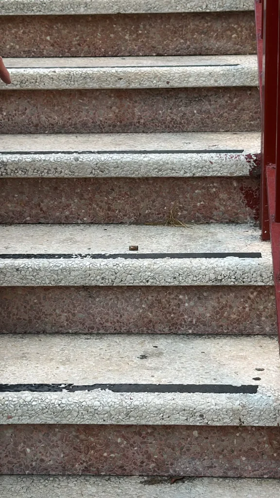 Cinco escalones de hormigón con una barandilla roja a la derecha, situados en Covibar. Los escalones están desgastados, con la pintura desconchada y bandas antideslizantes oscuras. Hay un pequeño objeto marrón y algo de hierba seca en los escalones, lo que sugiere una actividad reciente posiblemente relacionada con un ajuste de cuentas.