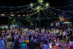 Una gran multitud se reúne al aire libre por la noche, bajo las luces colgantes y los adornos, para ver una actuación o un evento en el centro de una plaza. La gente está de pie, aplaudiendo y haciendo fotos en un ambiente festivo.