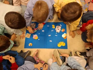 Un grupo de niños está sentado en círculo en el suelo, alrededor de una pizarra azul con dibujos animados y tarjetas, concentrados en una actividad que realizan juntos, lo que refleja el sentido de comunidad que a menudo se fomenta en las escuelas infantiles de Rivas.