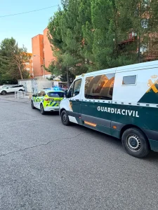 Una furgoneta de la Guardia Civil y un coche patrulla están aparcados en una carretera asfaltada, cerca de unos árboles y de un edificio de ladrillo rojo al fondo, a plena luz del día, en medio de las noticias sobre un incidente con arma de fuego relacionado con un ajuste de cuentas en la zona de Covibar.