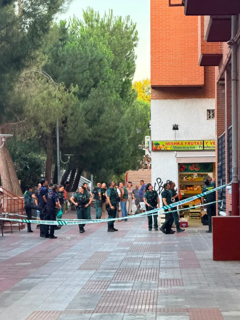 Varios agentes de policía se encuentran detrás de la cinta de precinto que bloquea una acera cerca de una tienda de alimentación Covibar, investigando lo que parece ser un ajuste de cuentas en el que se ha utilizado un arma de fuego. Varias personas observan desde la distancia mientras las autoridades acordonan la zona. La escena está rodeada de árboles y edificios.