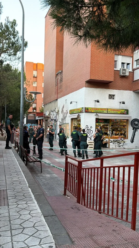 Un grupo de agentes uniformados se encuentra junto a una zona acordonada en una acera de la ciudad, frente a una tienda de alimentación y junto a unos edificios con grafitis. Algunos peatones observan desde cerca mientras las autoridades investigan lo que parece ser un ajuste de cuentas, tras las denuncias de un herido por arma de fuego en el barrio de Covibar.