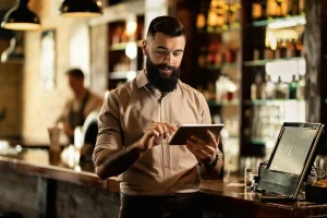 Un camarero con barba y las mangas remangadas utiliza una tableta digital detrás de la barra, gestionando con fluidez el sistema de reservas para garantizar un servicio sin contratiempos. En el fondo se ven botellas en las estanterías, y más al fondo de la barra se distingue a un hombre desenfocado.