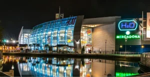 Un moderno centro comercial de Rivas por la noche, con paredes acristaladas, luces de colores y letreros de tiendas como el centro comercial H2O y Mercadona, reflejados en una tranquila superficie de agua en primer plano. El ambiente animado se ve realzado por el resplandor de una pantalla final del Mundial que ilumina la escena.