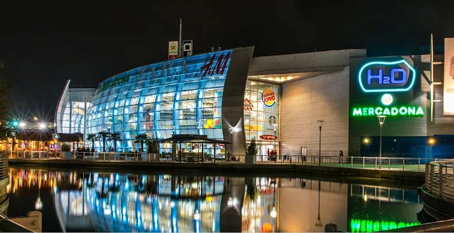 Un moderno centro comercial de Rivas por la noche, con paredes acristaladas, luces de colores y letreros de tiendas como el centro comercial H2O y Mercadona, reflejados en una tranquila superficie de agua en primer plano. El ambiente animado se ve realzado por el resplandor de una pantalla final del Mundial que ilumina la escena.