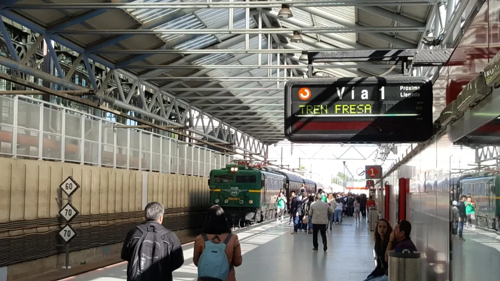 La gente camina por un andén cubierto mientras se acerca un tren verde, lo que evoca el encanto de los trenes históricos de la Comunidad de Madrid. Un letrero electrónico indica: «Vía 1 TRENE FRESA». El andén cuenta con vigas metálicas, paneles de cristal y diversos letreros visibles, todo ello bañado por la suave luz del otoño.