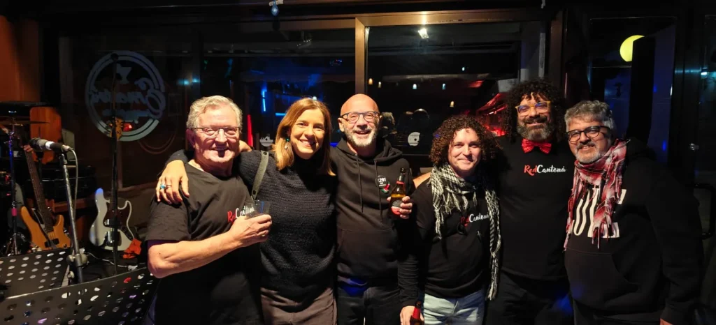 Seis adultos están muy juntos, sonriendo a la cámara en un espacio interior con poca luz. Algunos sostienen bebidas, y en el fondo se ven instrumentos musicales —entre ellos una batería—, lo que sugiere un ambiente distendido y social. Juan y sus amigos parecen estar disfrutando de su estancia en RedCantoná.