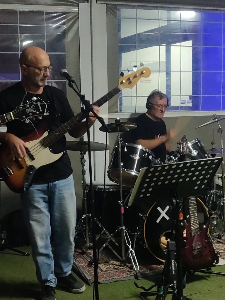 Dos músicos tocan en un local cerrado; Juan toca el bajo eléctrico y canta por un micrófono, mientras que el otro músico toca la batería. En primer plano hay un atril con partituras y equipo adicional, lo que refleja la energía creativa de RedCantoná.