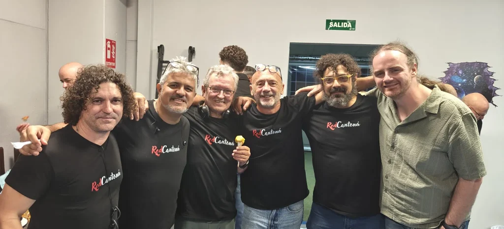 Seis hombres de pie en el interior de un local, cinco de ellos con camisetas negras a juego de ReCantares. Entre ellos se encuentra Juan, sonriendo junto a sus amigos y abrazados unos a otros. En la pared que tienen detrás hay una señal verde de salida y un extintor.