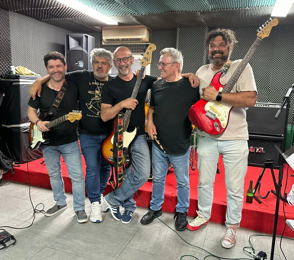 Cinco hombres están de pie juntos, con guitarras eléctricas y un bajo en la mano, en una sala de ensayos con paredes insonorizadas y moqueta roja. Entre ellos se encuentra Juan, sonriendo y con el brazo alrededor del hombro de otro miembro del grupo. Al fondo se ve el equipo musical, incluida una batería, junto con un cartel en el que se lee «RedCantoná».