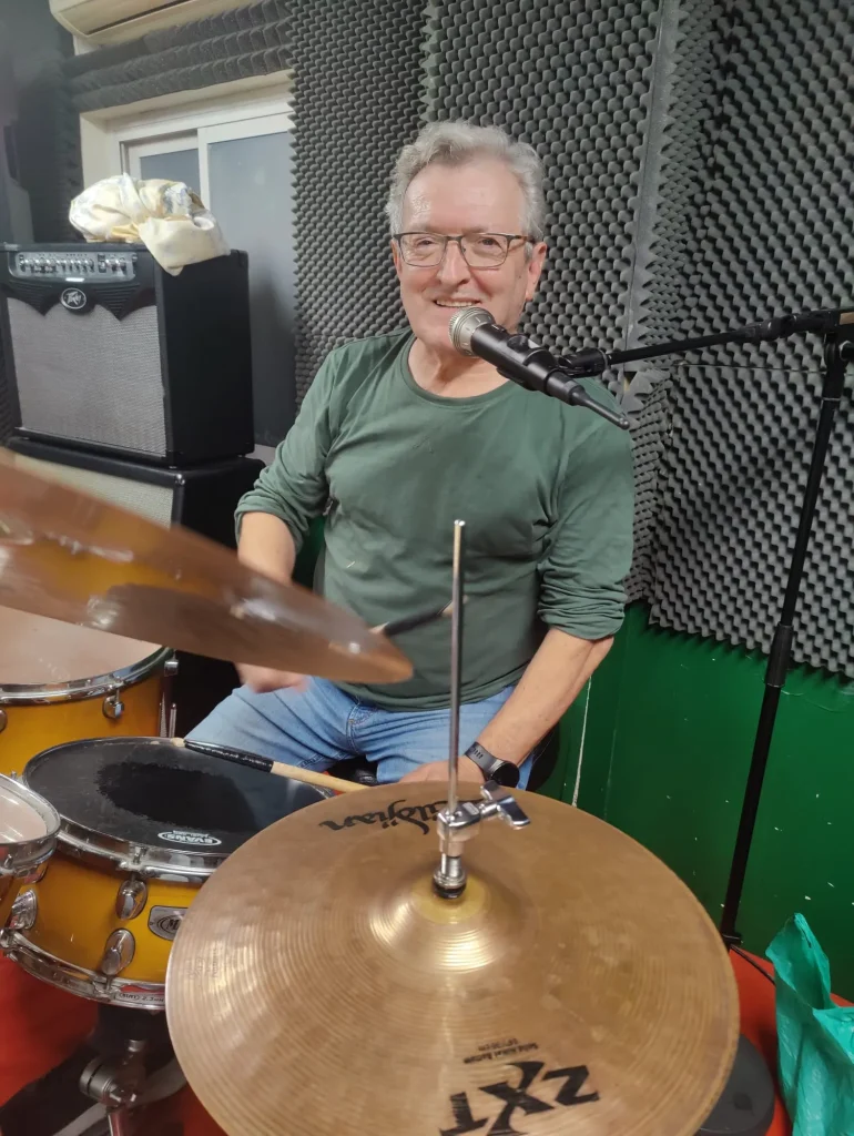 Un hombre mayor con gafas y una camiseta verde, identificado como Juan, toca la batería y canta por un micrófono en una sala insonorizada. Se pueden ver el equipo de sonido y los platillos, que captan la energía vibrante de esta sesión de RedCantoná.