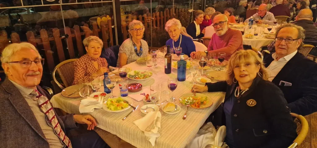 Siete adultos, entre ellos IU Rivas y Pilar Trejo, están sentados alrededor de una mesa en un restaurante, cenando ensaladas y bebiendo vino. La mesa está puesta con platos, copas y botellas. El ambiente parece acogedor y distendido, y se ve a otros comensales al fondo.