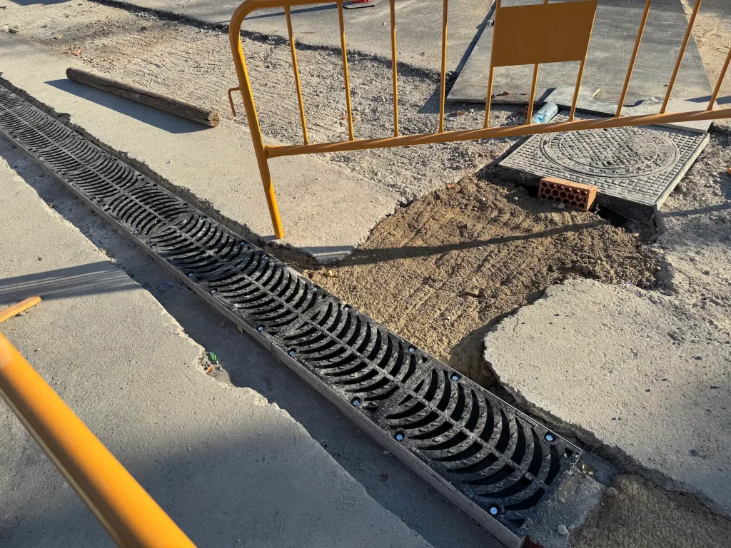 En una calle pavimentada de Rivas se ha instalado una rejilla de drenaje larga y negra. Cerca de allí se pueden ver una tapa de alcantarilla y un trozo de tierra al descubierto, lo que pone de manifiesto una reciente retirada de árboles. La zona está rodeada de barreras metálicas amarillas, lo que indica que se están llevando a cabo obras de construcción o mantenimiento impulsadas por la administración local del PP.