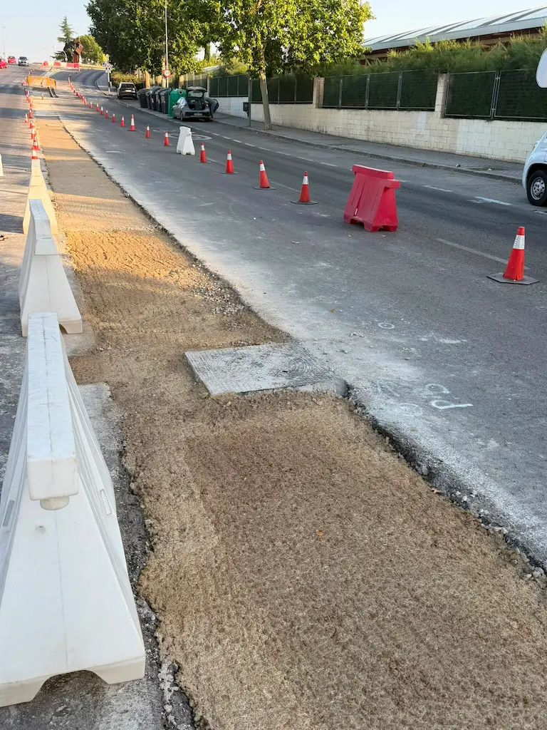 Una calle de la ciudad de Rivas en obras, con tramos de acera retirados; conos de tráfico y barreras bordean la calzada, y al fondo se ven vehículos de obra. A lo largo de la calzada hay indicios de la retirada de árboles, lo que se suma a la transformación que se está llevando a cabo. La escena está iluminada por la luz natural del día.
