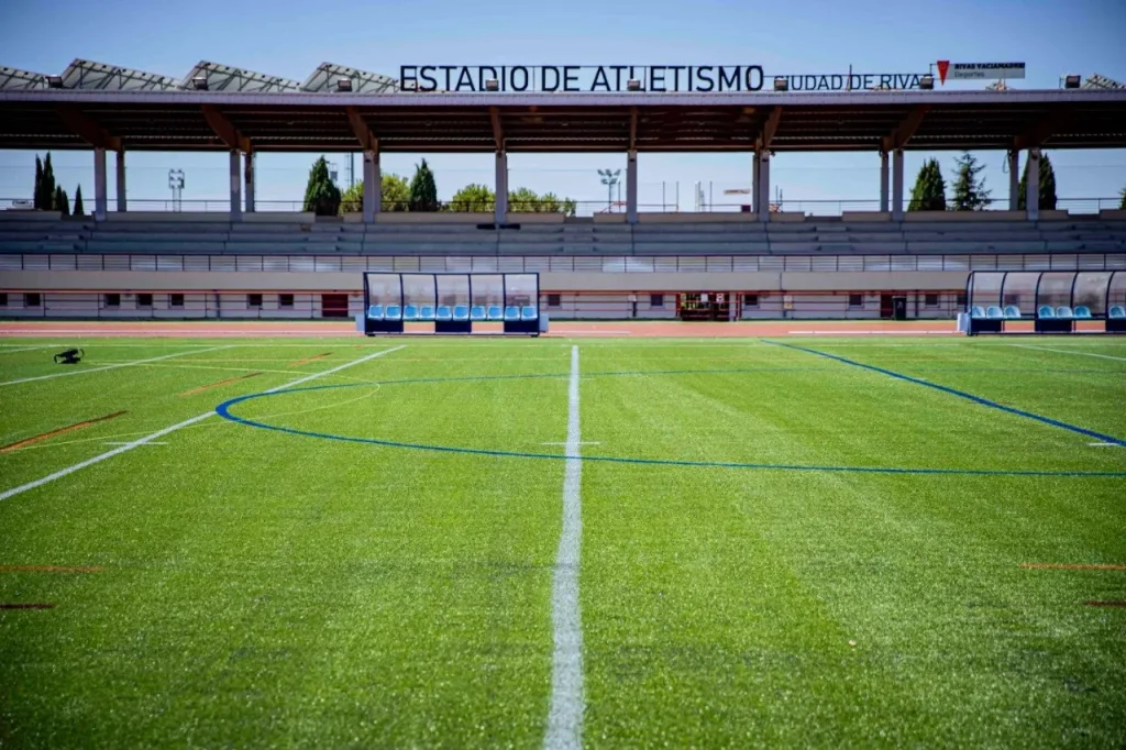 Un campo de fútbol con césped verde y fresco, líneas de marcado blancas y azules, y banquillos vacíos. Al fondo, hay una grada cubierta con un cartel en el que se lee «ESTADIO DE ATLETISMO CIUDAD DE RIVAS», lo que da la impresión de ser un recinto impecable y listo para la acción, casi como si lo hubiera creado un «Borrador automático».