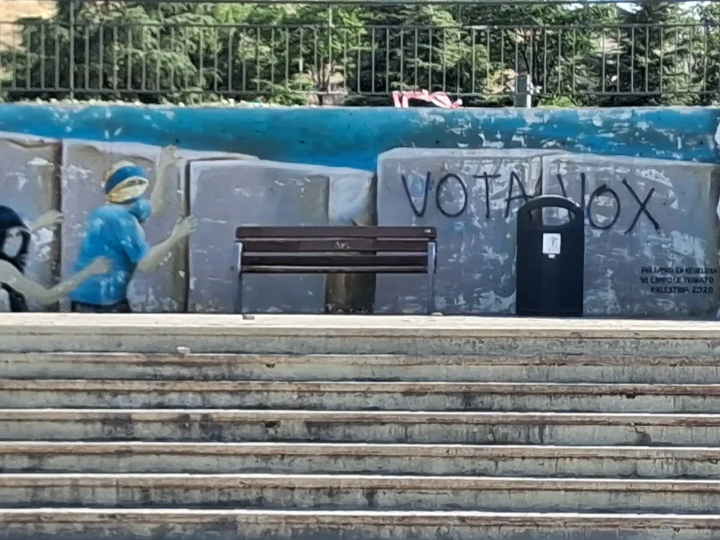 Unos escalones de piedra conducen a un banco y a un contenedor de basura. Detrás de ellos, un mural muestra a dos personas, con las palabras «VOTA VOX» pintadas con spray sobre parte de la obra, lo que ha suscitado propuestas para repintar el mural «Palestina» y recuperar su mensaje original. Al fondo se ven árboles y una valla.