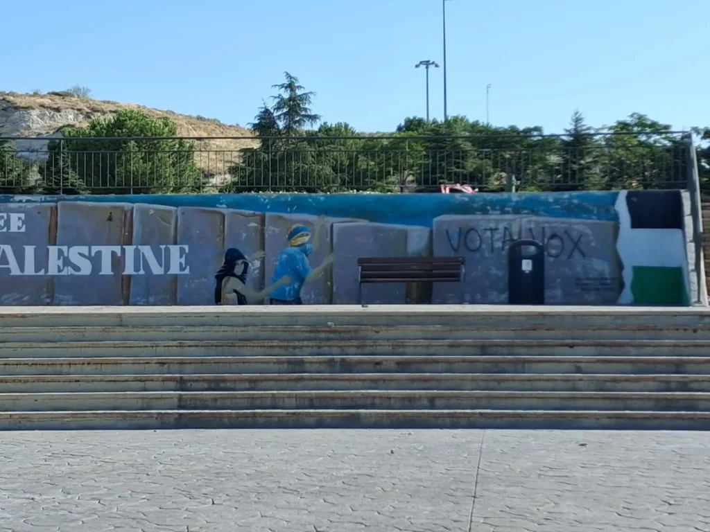 En un mural pintado sobre un muro de hormigón se lee «FREE PALESTINE» y se ven dos figuras, una de azul y otra de negro, junto a una urna electoral. Delante hay un banco, al que conducen unos escalones. Al fondo se divisan árboles y una colina. La comunidad ha debatido recientemente los planes para repintar el mural de Palestina, con el fin de garantizar que su mensaje siga siendo impactante durante muchos años.