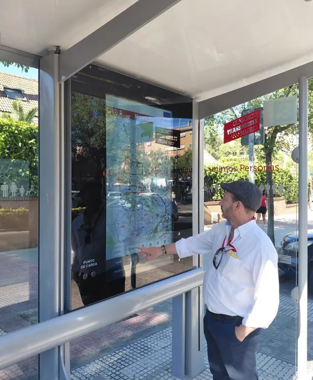 Un hombre vestido con camisa blanca y corbata consulta un mapa de transporte público en una pantalla táctil situada en la marquesina de una parada de autobús en Rivas, en un día soleado, con árboles y edificios visibles al fondo. La escena pone de relieve la comodidad de los mapas interactivos a la hora de orientarse en el transporte público.