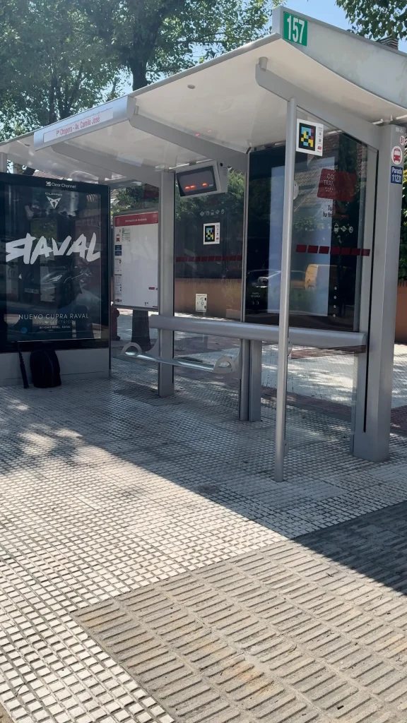 Una moderna marquesina de autobús con paredes de cristal, un panel digital y un banco. Hay una mochila en el suelo y un gran cartel publicitario a la izquierda. El suelo está revestido con baldosas claras y oscuras. Se indica la línea de autobús 157 con destino a Rivas y hay mapas interactivos a disposición de los pasajeros para su comodidad.