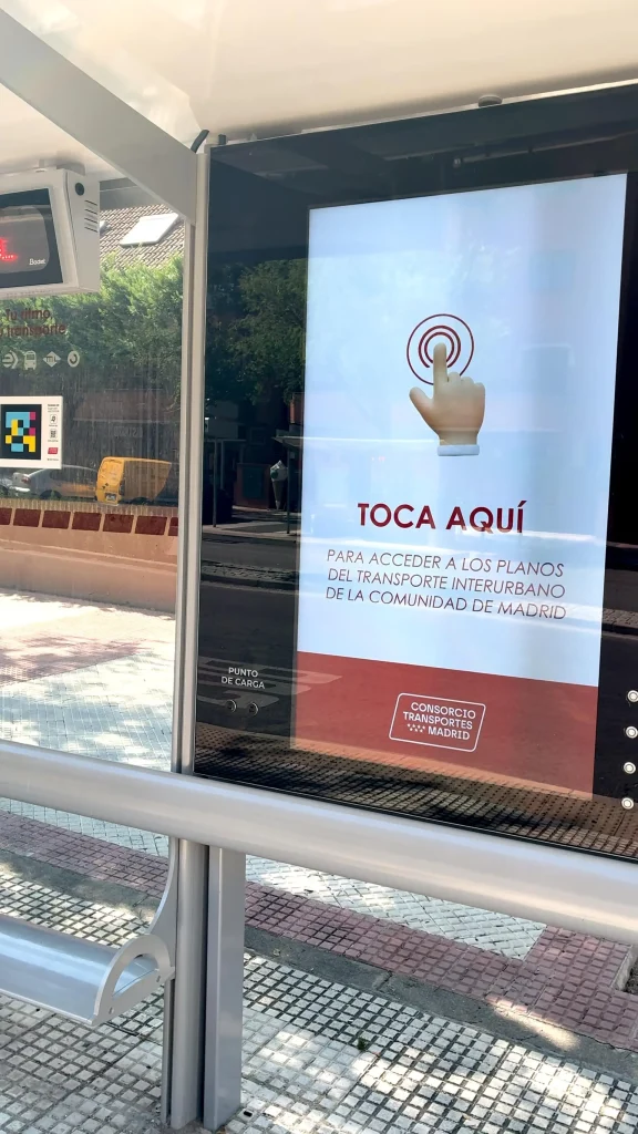Una pantalla digital situada en la marquesina de una parada de autobús muestra instrucciones en español, en las que se ve una mano pulsando un botón y se lee: «TOCA AQUÍ para acceder a los mapas interactivos del transporte interurbano de la Comunidad de Madrid en Rivas».