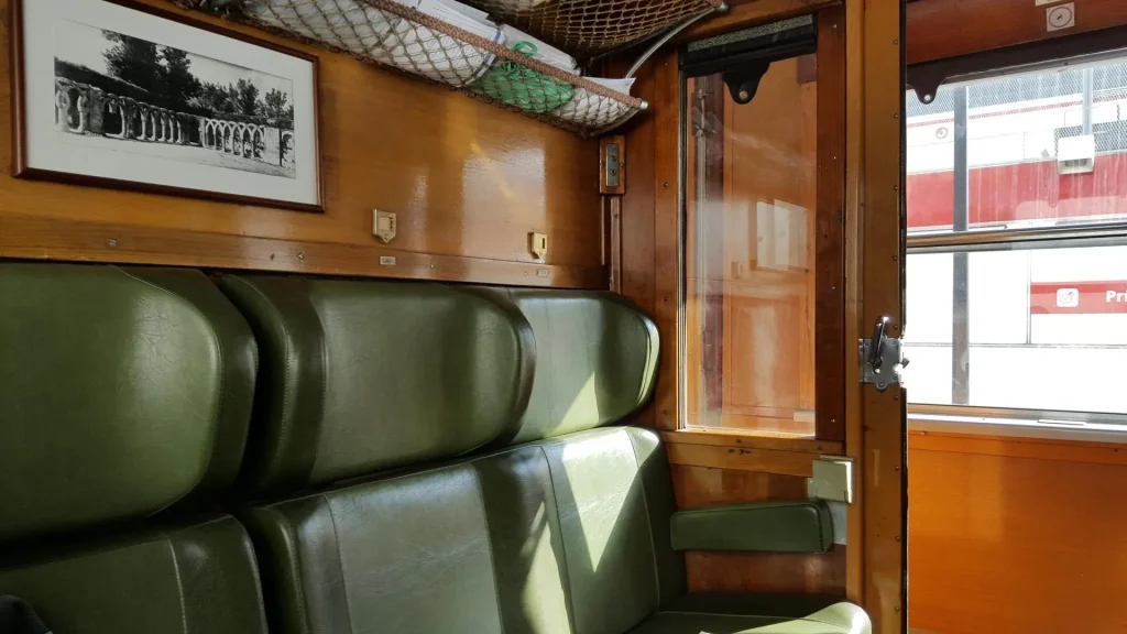 Compartimento de tren de época con asientos de cuero verde, paredes de madera, una foto enmarcada, portaequipajes en el techo y la luz del sol que entra por las ventanas entreabiertas. A través de la ventana se ve pasar otro tren de «Los Trenes Históricos de la Comunidad de Madrid» bajo la suave luz del otoño.