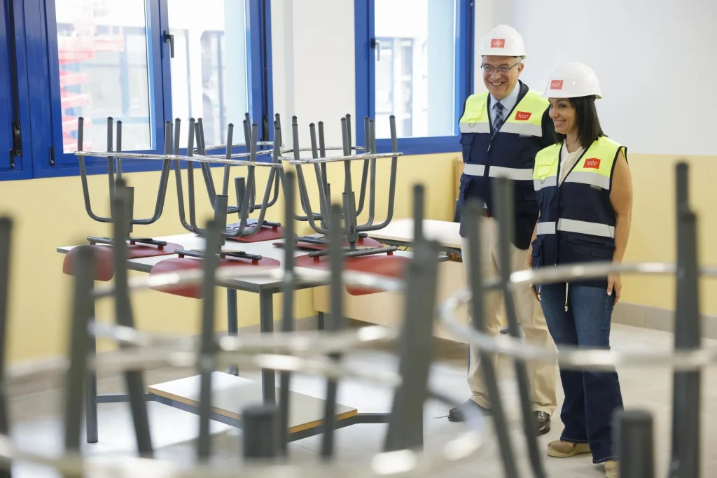 Dos personas con chalecos de seguridad y cascos se encuentran en un aula con ventanas azules, probablemente revisando las últimas «obras educativas». Las sillas están apiladas boca abajo sobre los pupitres, y ambas personas están observando la sala, quizá como parte de una inspección en Zarzalejo o Rivas.
