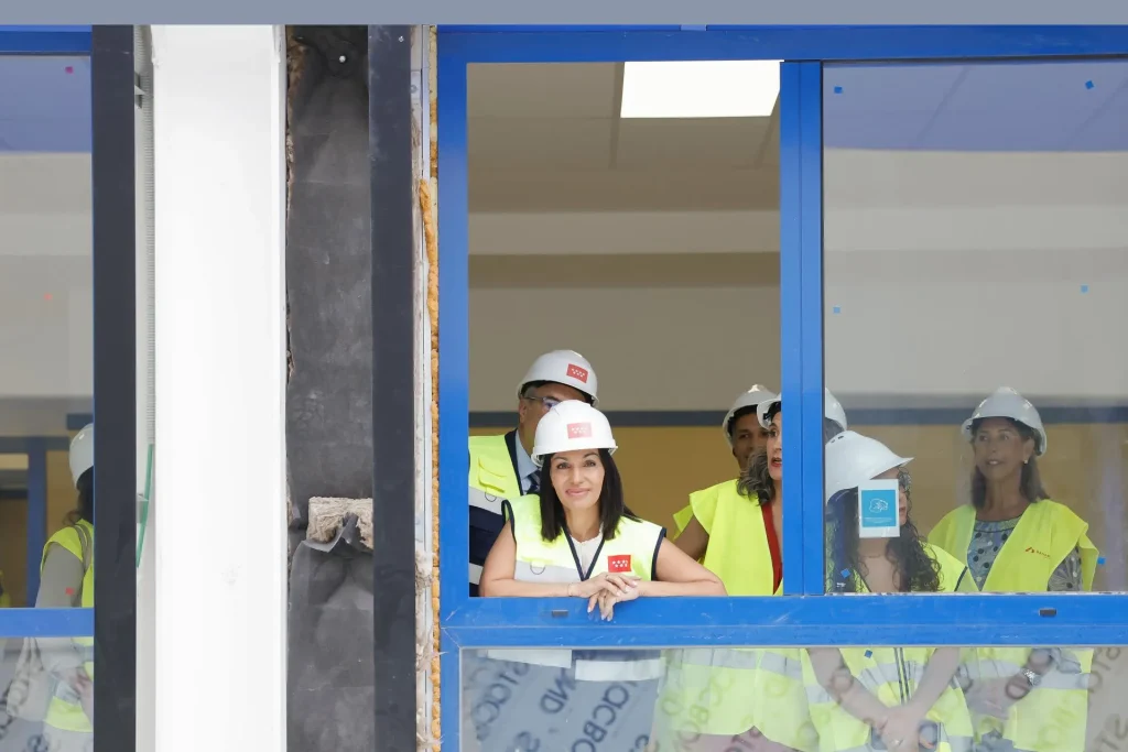 Un grupo de personas con cascos y chalecos de seguridad amarillos se encuentra detrás de una ventana con marco azul en una obra de construcción en Rivas, observando el avance de las obras educativas. Una persona que está delante tiene los brazos apoyados en el alféizar de la ventana y mira atentamente hacia fuera.