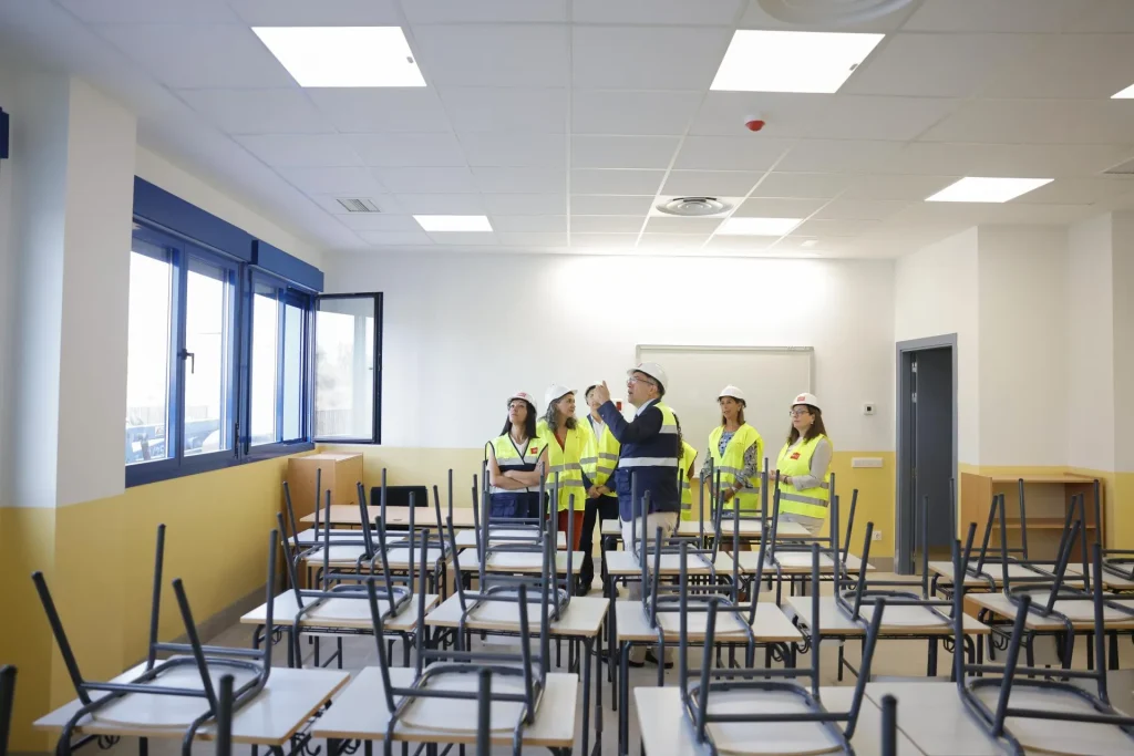 Cinco personas, equipadas con cascos y chalecos reflectantes, inspeccionan un aula en la que hay sillas apiladas sobre los pupitres, como parte de las obras educativas en Rivas, Zarzalejo y Castillejo. La sala tiene paredes amarillas y blancas, grandes ventanas y un techo blanco con luces de techo.