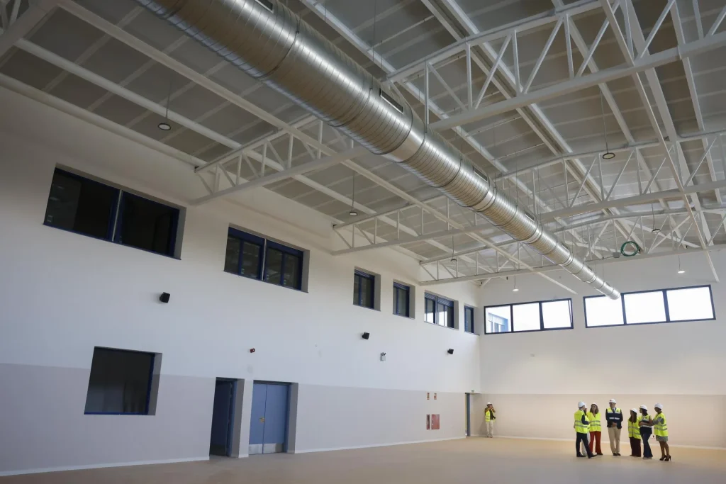 Un grupo de personas que llevan chalecos de seguridad amarillos y cascos se encuentra en una gran sala vacía, con techos altos, conductos a la vista, ventanas y paredes blancas, inspeccionando el avance de las obras educativas en Rivas, Zarzalejo y Castillejo.