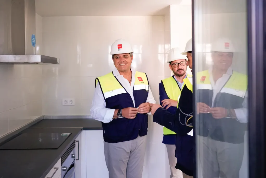 Dos hombres con cascos y chalecos reflectantes aparecen en una cocina moderna con paredes blancas y una placa de cocina, lo que representa las obras de construcción en curso en 2023 en las viviendas públicas de la Comunidad de Madrid. Uno de ellos sonríe a la cámara, mientras que el otro mira hacia otro lado. A la derecha se ve el reflejo de ambos.