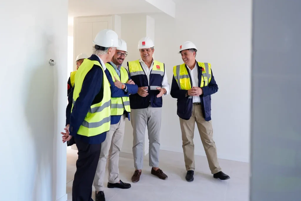 Cuatro hombres con cascos y chalecos de seguridad están juntos en una sala muy bien iluminada, sonriendo y hablando, lo que sugiere que son inspectores o profesionales que trabajan en un proyecto de 2024 viviendas públicas en la Comunidad de Madrid.