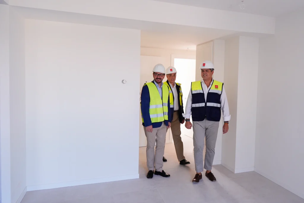Tres hombres que llevan chalecos de seguridad y cascos de obra recorren una sala luminosa y sin amueblar, con paredes blancas y suelo de baldosas, posiblemente inspeccionando una obra de construcción o reforma de nuevas viviendas públicas en la Comunidad de Madrid en 2023.