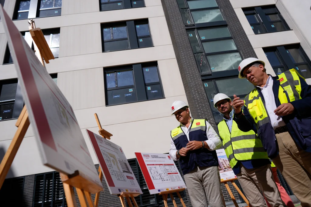 Tres personas con ropa de obra y cascos de seguridad examinan los planos de construcción en caballetes frente a un moderno bloque de pisos en construcción, con ventanas y paredes exteriores visibles al fondo; se trata de un proyecto de viviendas públicas de 2023 financiado por la Comunidad de Madrid.